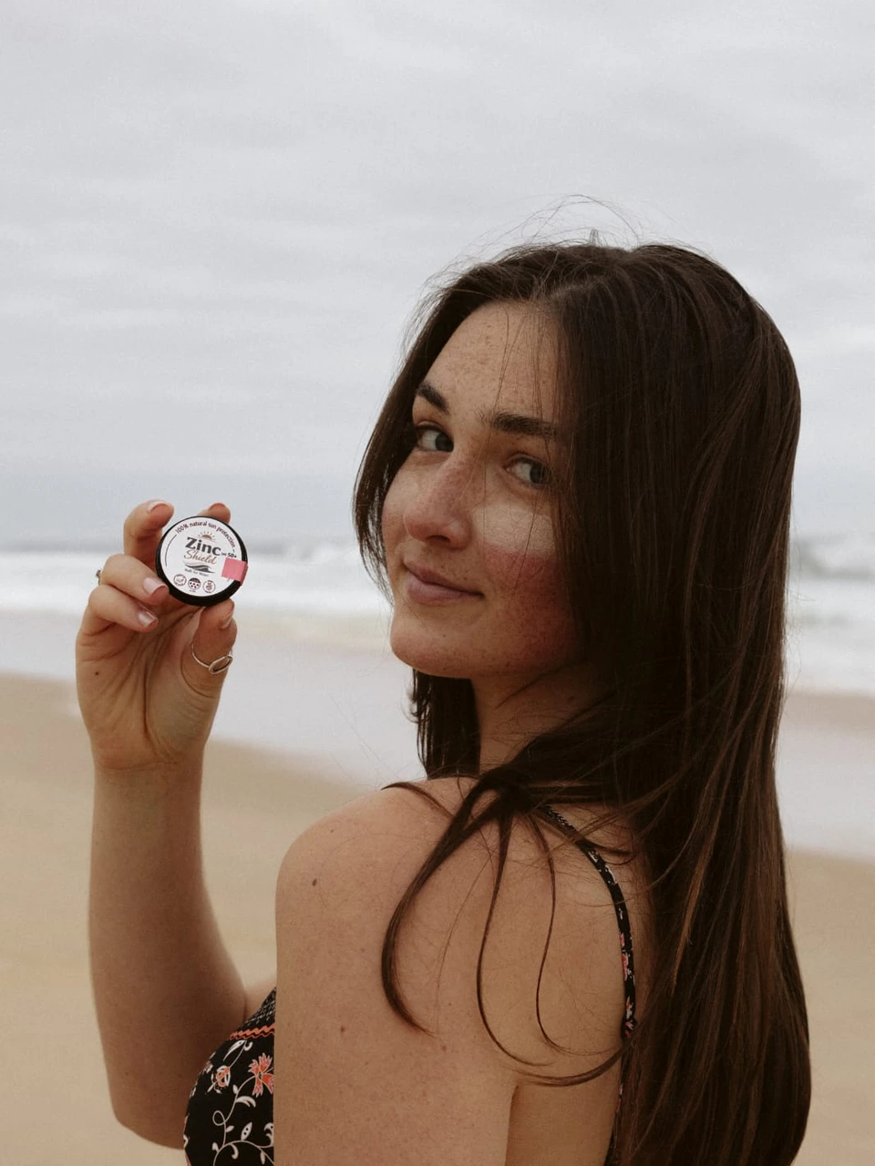 Woman holding ZincShield SPF 50 mineral sunscreen tin on the beach.